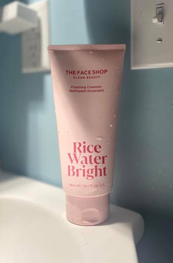 A tube of The Face Shop Clean Beauty Rice Water Bright foaming cleanser with water droplets on it, placed on a bathroom counter