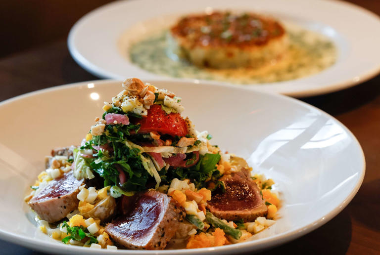 A Pearl Niçoise salad (front) sits in front of The Crab Cake at Bobbie's Airway Grill in the Preston Royal shopping center in Dallas on Tuesday, May 30, 2023. The salad is made with seared ahi tuna, new potatoes, green beans, Kalamata Olives, tomatoes and almonds.