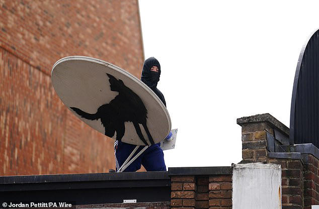 A hooded man with a mask is seen holding a satellite dish with Banksy's wolf mural on it before the work disappeared