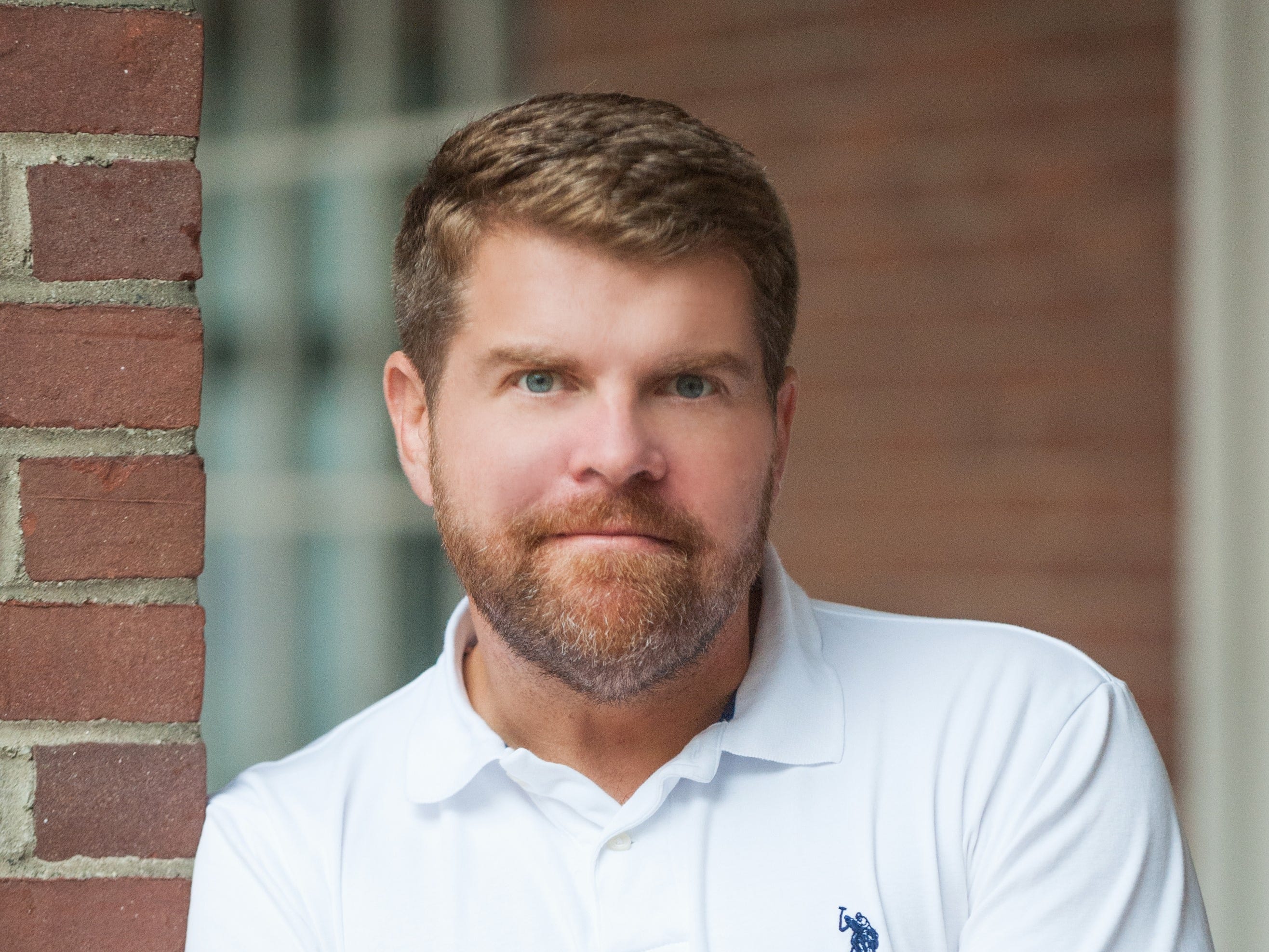 A man (former FBI agent Jason Hogg) stands wearing a white polo shirt with arms folded against a brick wall.