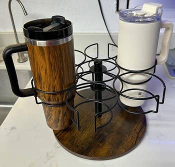 Two travel mugs, wood-patterned and white, placed on a rotating rack in a kitchen setting. The rack can hold up to six mugs and is on a marble countertop