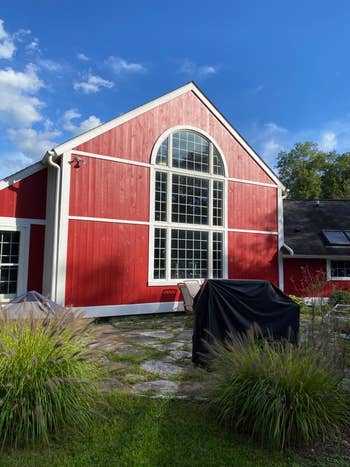 A red barn-style house with large arched windows and a patio featuring covered outdoor furniture. Green plants frame the foreground