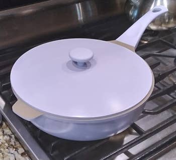 A covered frying pan sits on a stove next to a wooden spatula and a metal spoon rest in a kitchen setting
