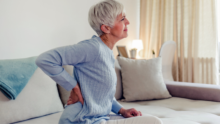 woman sitting on couch holding lower back in discomfort due to one of the top causes of sciatica