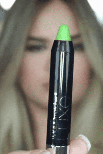 Close-up of a person holding a green-tipped makeup pencil in front of a blurred background