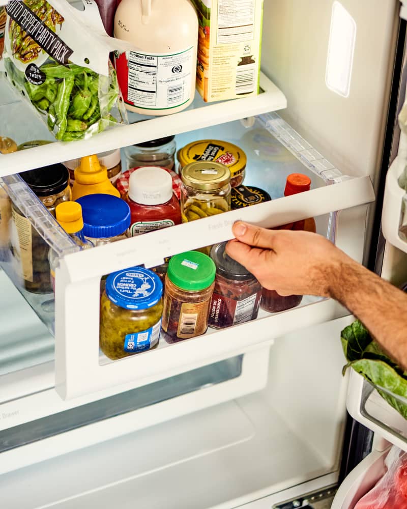 Hand pulling out a drawer/bin in a refrigerator. The bin is full of condiments (catsup, mustard, hot sauce, etc)