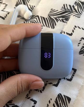 Hand holding wireless earbuds case with a digital display showing battery level
