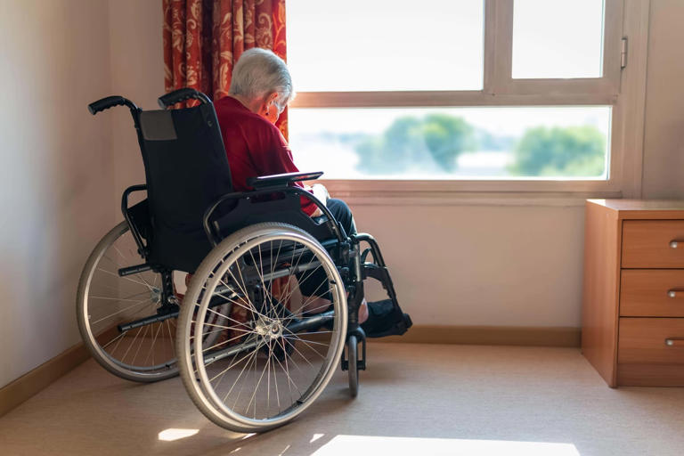 An older woman with white hair, alone in her house while looking outside through the window waiting for company