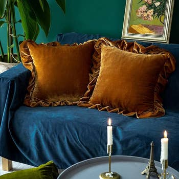 Stylish living room with saffron velvet pillow covers on a blue sofa, flanked by a plant, with candles and artwork creating a cozy, elegant ambiance
