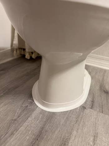 Bottom view of a white toilet, showing its base against a wooden floor. The image highlights the toilet's installation and design