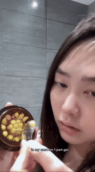 Person applying cream with a scoop to their face from a small round container, demonstrating skincare product application