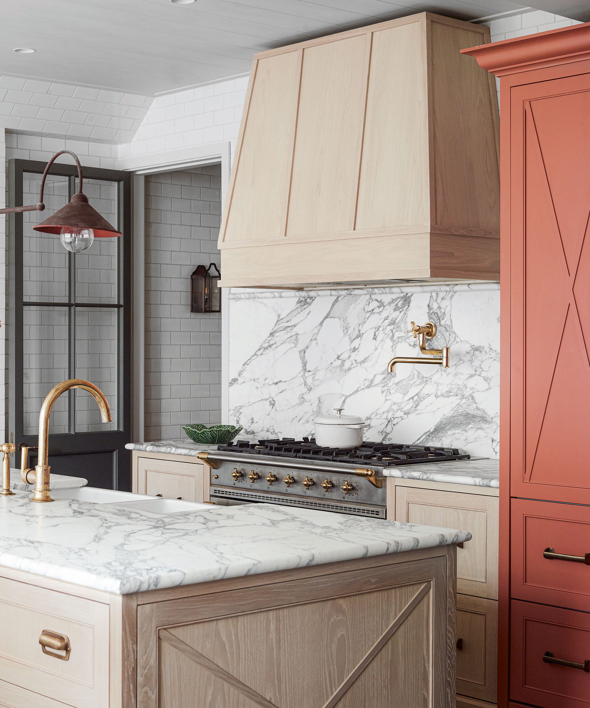 kitchen with island, stove and marble surfaces and backsplashes. There is a large range, hood, and feature terracotta colored tall cabinet whilst the others are pale wood