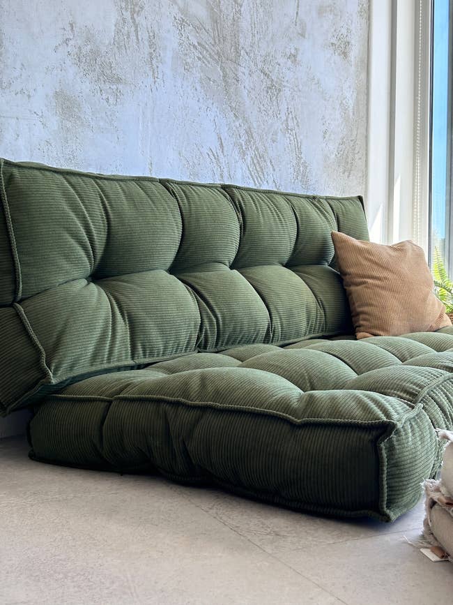 Tufted green sofa with a tan pillow next to a window, set against a textured wall, creating a cozy seating area