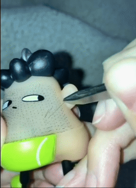 Toy figure with black hair and a green belt being sculpted with a tool, creating texture on its cheek