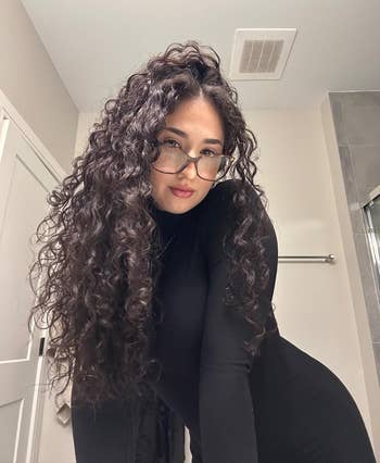 reviewer with long curly hair wearing glasses and a black turtleneck dress poses indoors