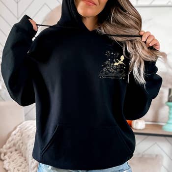 Model models a black hoodie with a subtle star and plant design, pairing it with casual denim shorts, suitable for a relaxed shopping style