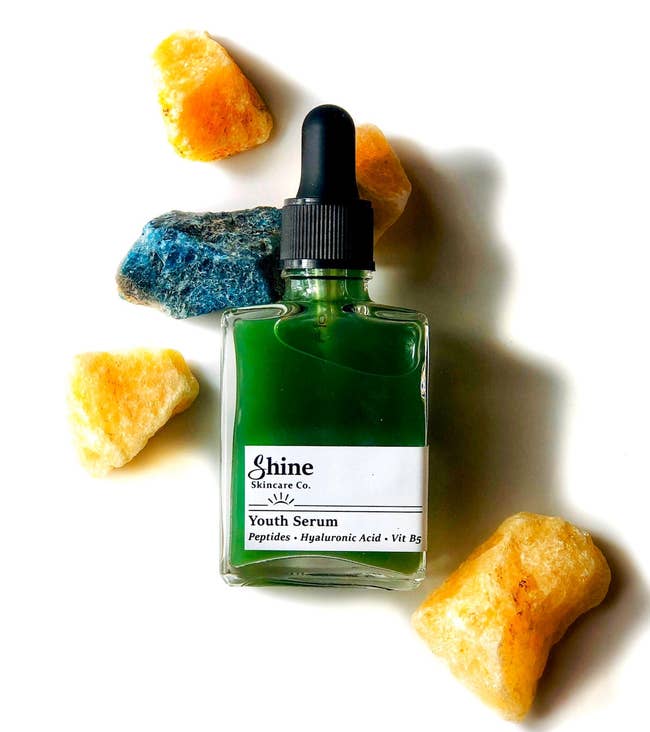 A bottle of green youth serum by Shine Skincare Co., surrounded by colorful rocks, highlighting skincare product focus for a shopping feature