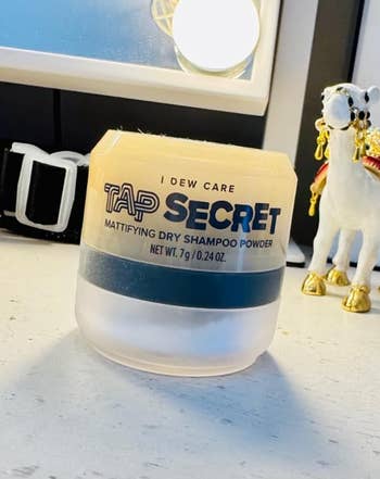 I Dew Care Tap Secret mattifying dry shampoo powder jar on a vanity, with a decorative horse figurine nearby