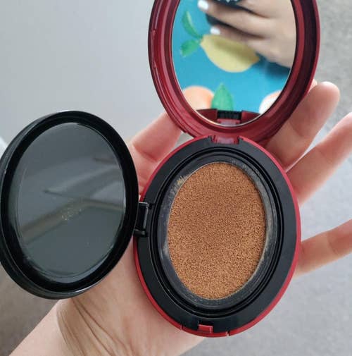 A hand holding an open compact foundation with a cushion makeup sponge. In the background, another hand with painted nails holding what appears to be a phone or notebook