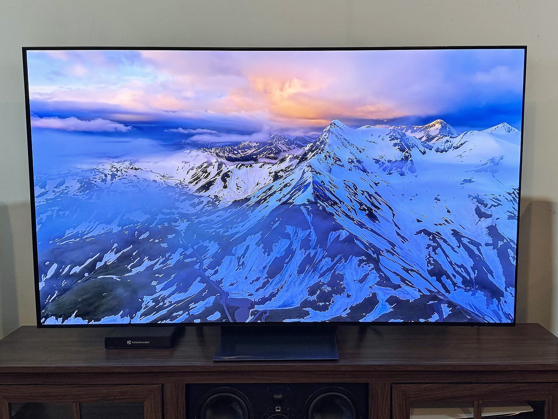 A Samsung S90F TV is on a media console, and the screen displays an image of a snowy mountain range.