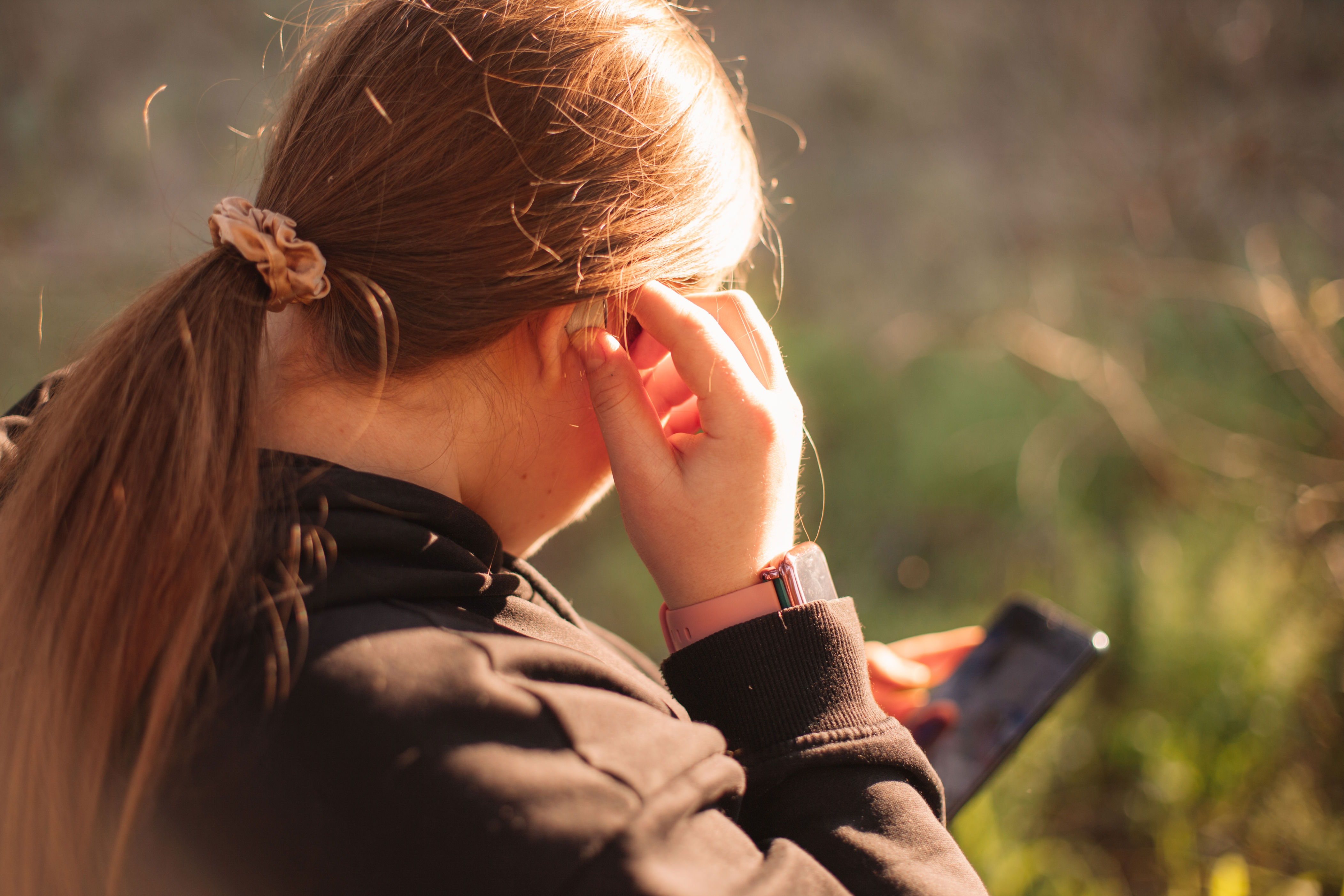 Person with a ponytail and smartwatch looks at smartphone outdoors, wearing a hoodie