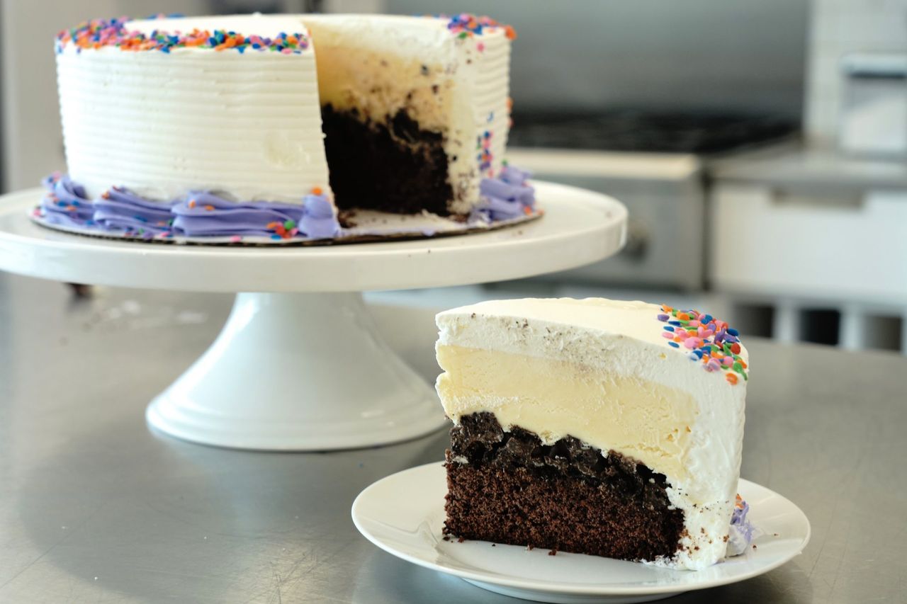 side view of a slice of homemade ice cream cake with a chocolate cake base, crunchy cookie filling, vanilla ice cream top layer, and whipped cream frosting with candy sprinkles