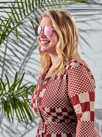Person smiling, wearing stylish geometric-patterned outfit and reflective sunglasses, posing near a leafy plant