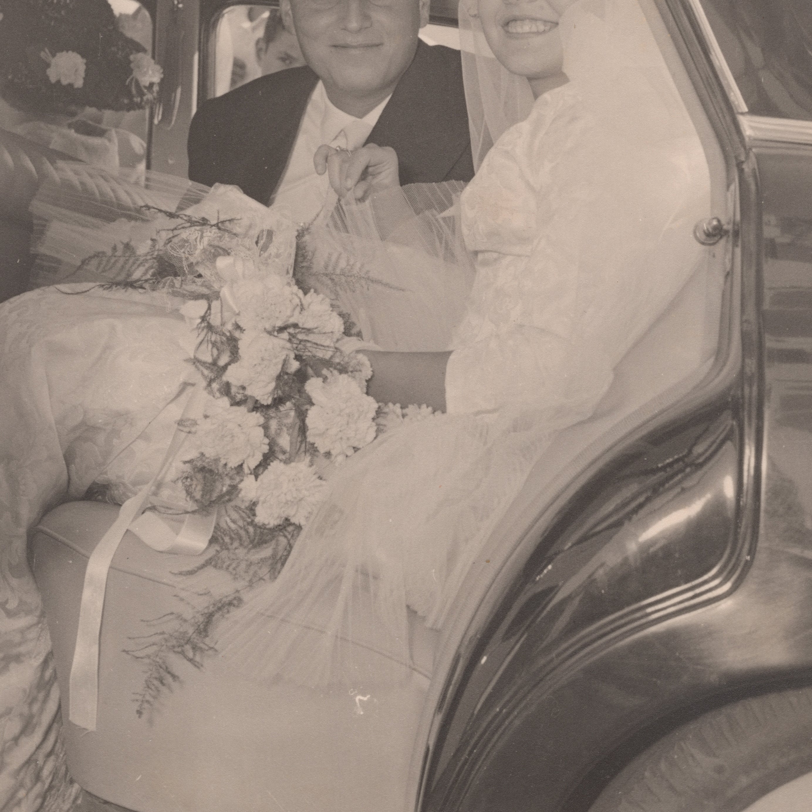 A couple in wedding attire smiles while seated in the back of a car, with the bride holding a bouquet and wearing a veil