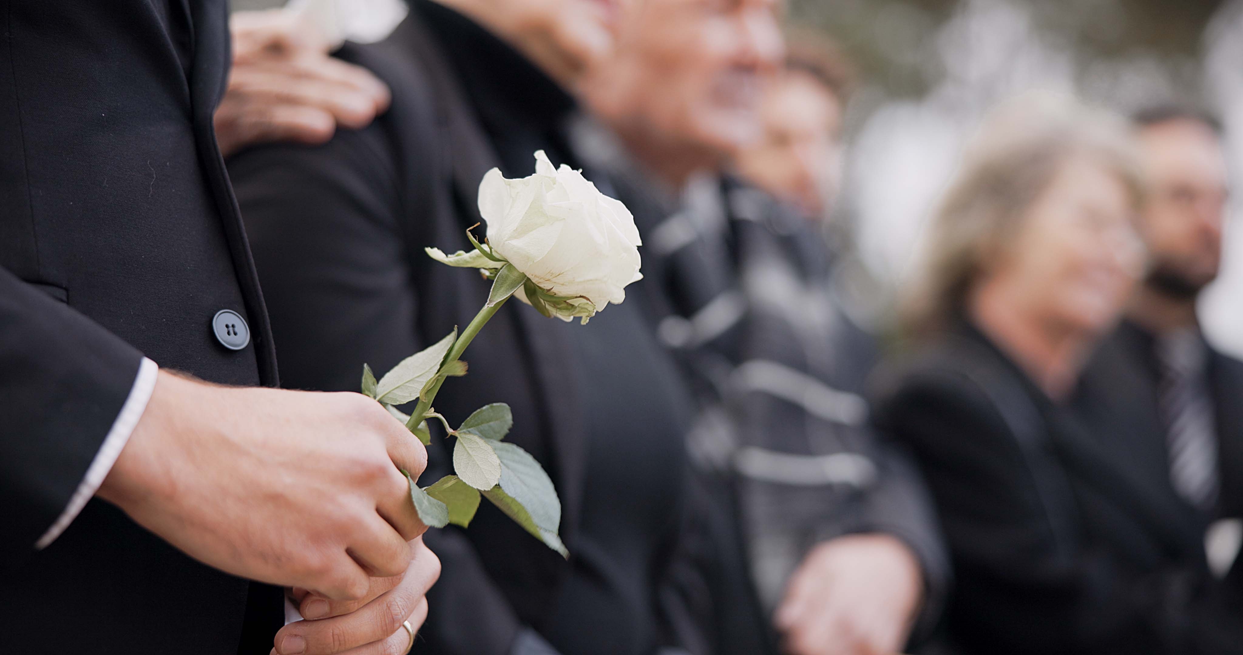 Person holding a white rose at a gathering. Others stand nearby, dressed formally, suggesting a somber event