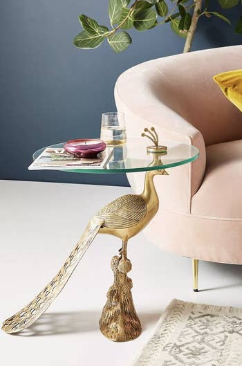 Elegant peacock-themed side table with glass top next to plush chair, featuring a plant in the background. Great for adding style to living spaces