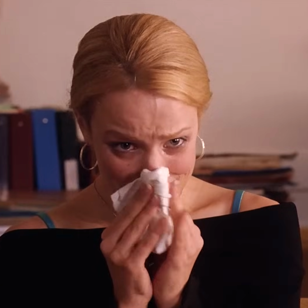 A person sitting indoors, looking concerned, holds a tissue to their nose, appearing as though they might be crying or experiencing allergies