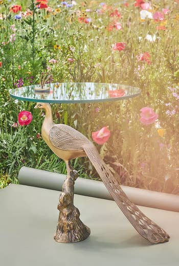 Glass-top table with a peacock sculpture base in a garden setting with vibrant flowers. Ideal for outdoor decor