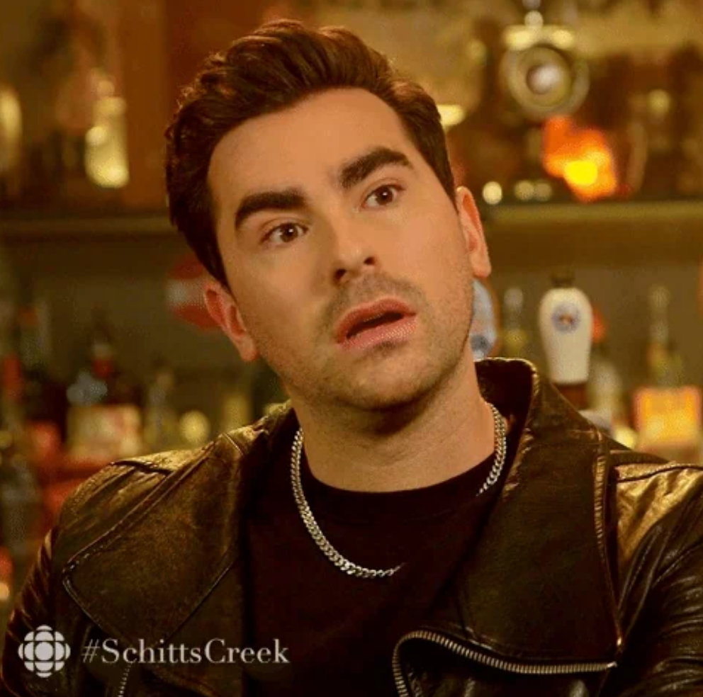A person with a surprised expression, wearing a black leather jacket and chain necklace, in a bar setting. Hashtag Schitt's Creek visible