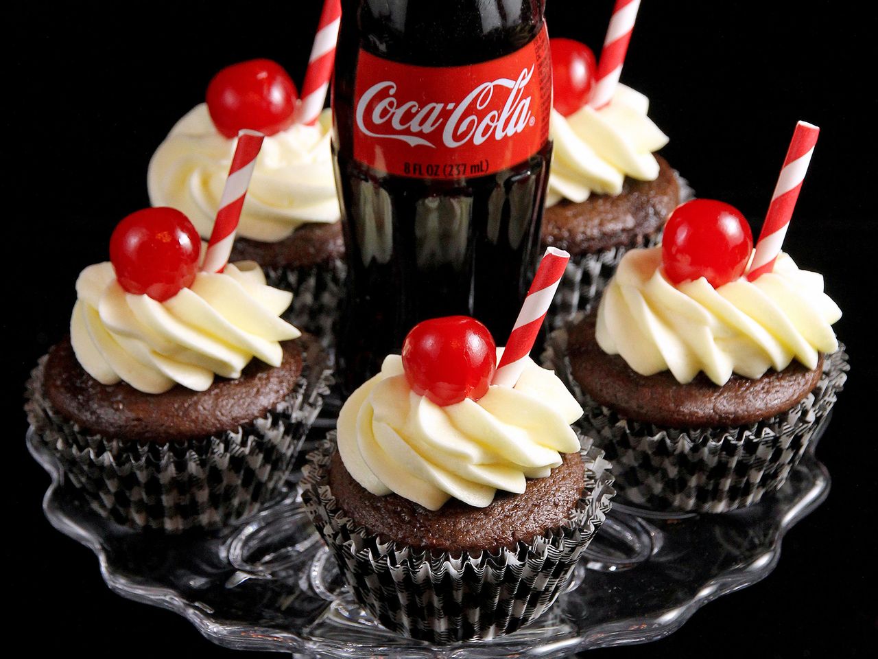 close up view of Cola cupcakes garnished with icing, cherries and straws and a coca cola bottle on a cake stand