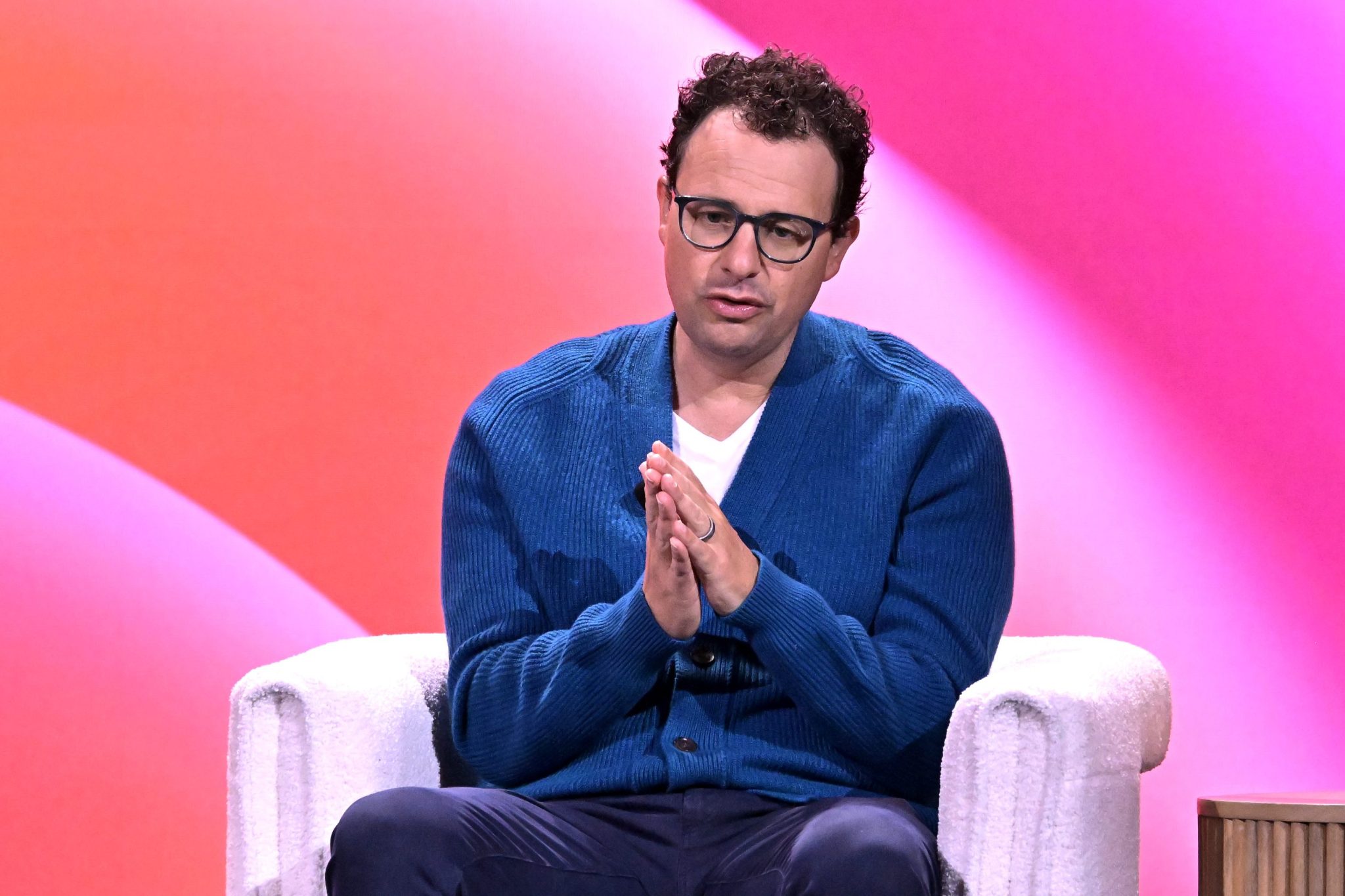 Dario Amodei sits in a white chair with his hands pressed together in front of a pink and orange background.