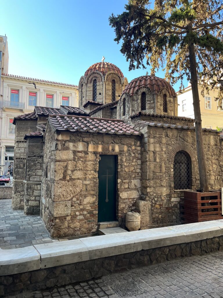 Small historic stone church with red tiled domes and a green door sits between modern buildings on a quiet street in Athens.
