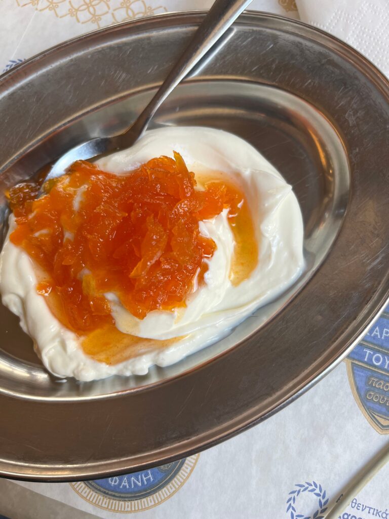 Thick Greek yogurt topped with bright orange fruit preserves sits in a metal dish with a spoon on the side.