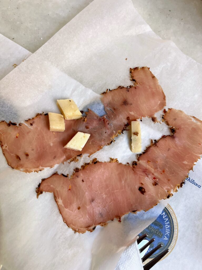 Thin slices of cured meat topped with small cubes of cheese are arranged on white paper on a table.