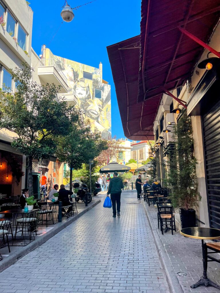Lively pedestrian street in Athens lined with cafes and outdoor seating as people walk under a clear blue sky with a mural on a nearby building capturing the atmosphere when planning how many days in Athens.