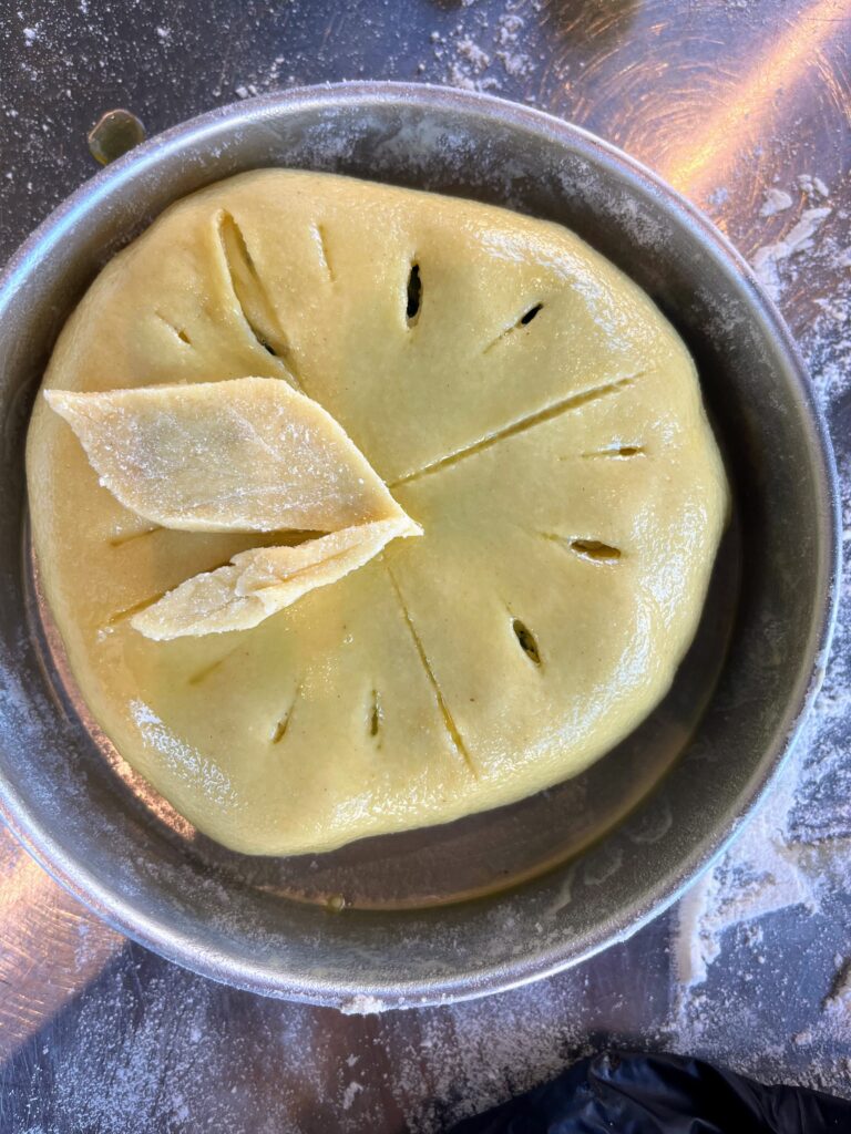 Round dough scored on top with a decorative leaf detail rests in a metal pan on a floured surface before baking.