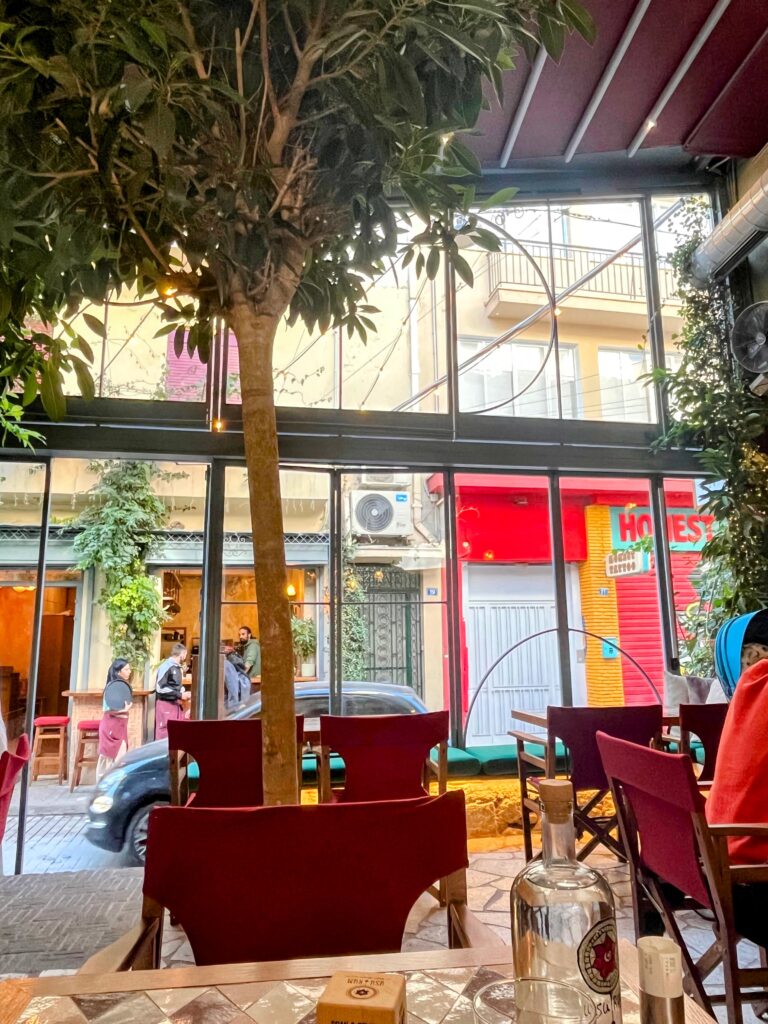 Cozy cafe interior with red chairs and a tree growing inside beside large windows looking out onto a narrow street with people and colorful storefronts.