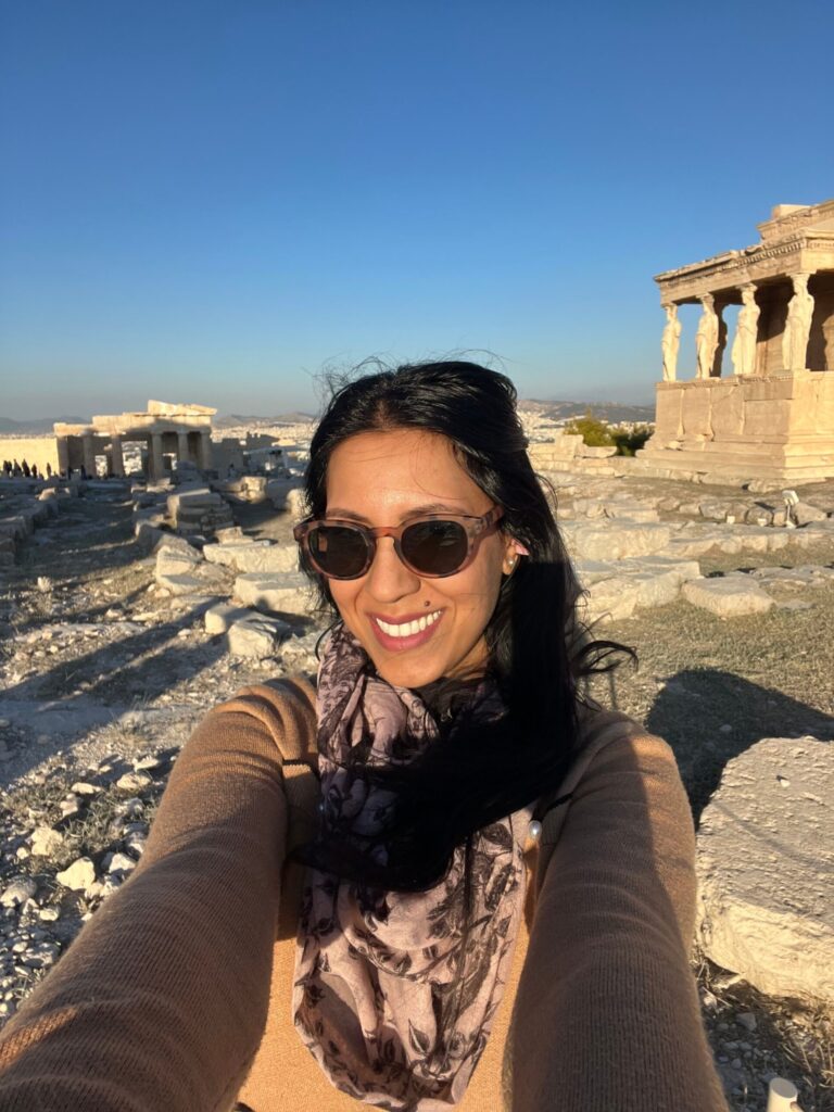 Woman wearing sunglasses smiles at the camera while standing among ancient ruins in Athens with stone columns and a clear blue sky behind her.