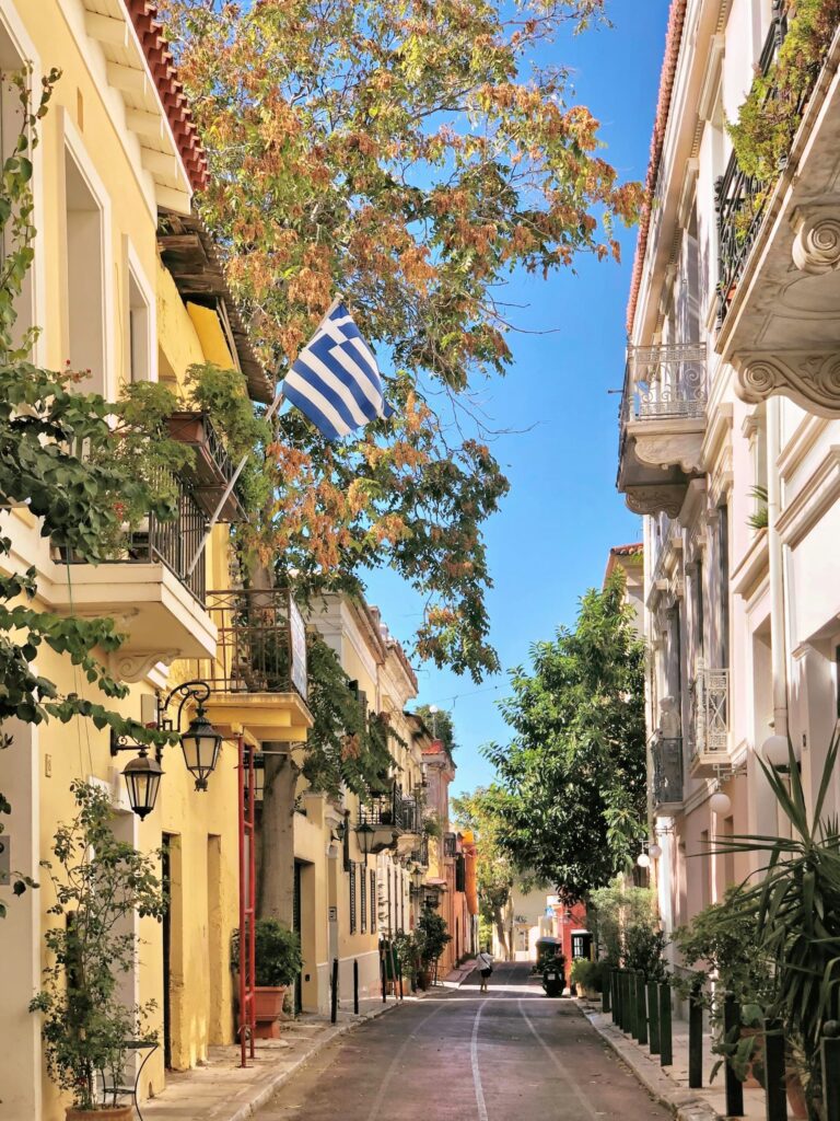 Quiet narrow street in Athens lined with pastel buildings balconies and plants with a Greek flag hanging above the road under a bright blue sky.