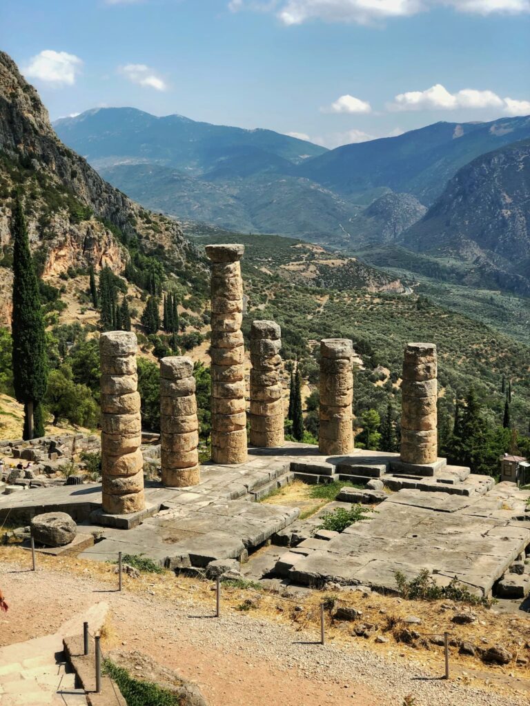 Ancient stone columns at Delphi overlook a wide mountain valley with green hills and winding roads stretching into the distance.