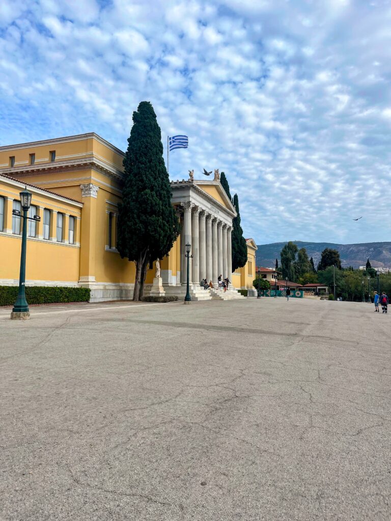 Neoclassical yellow building with tall columns and a Greek flag flying above it in Athens with trees and an open plaza in front.