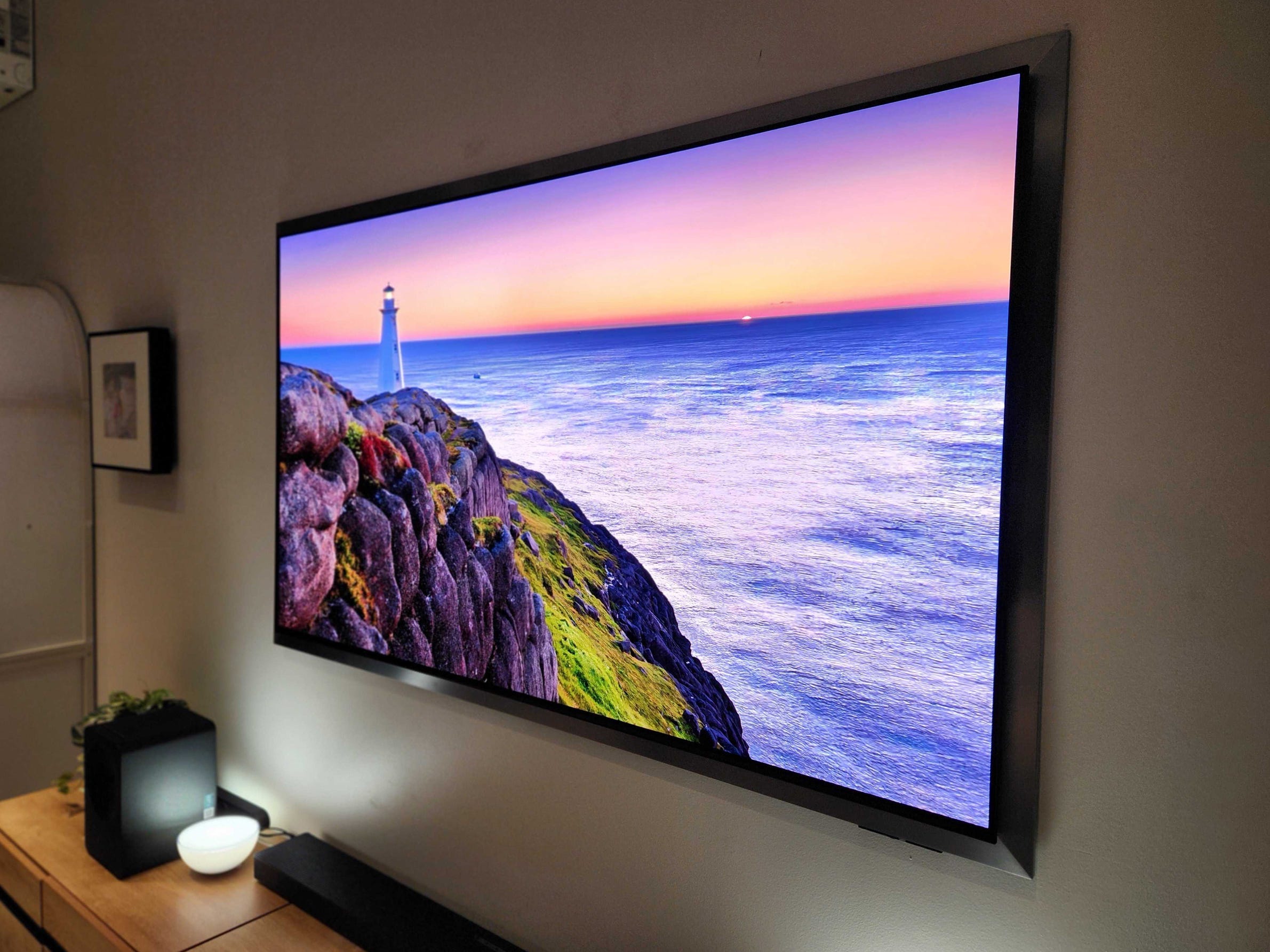 An angled view of a Samsung S95H OLED TV hanging on a wall, displaying an image of a lighthouse overlooking the ocean.