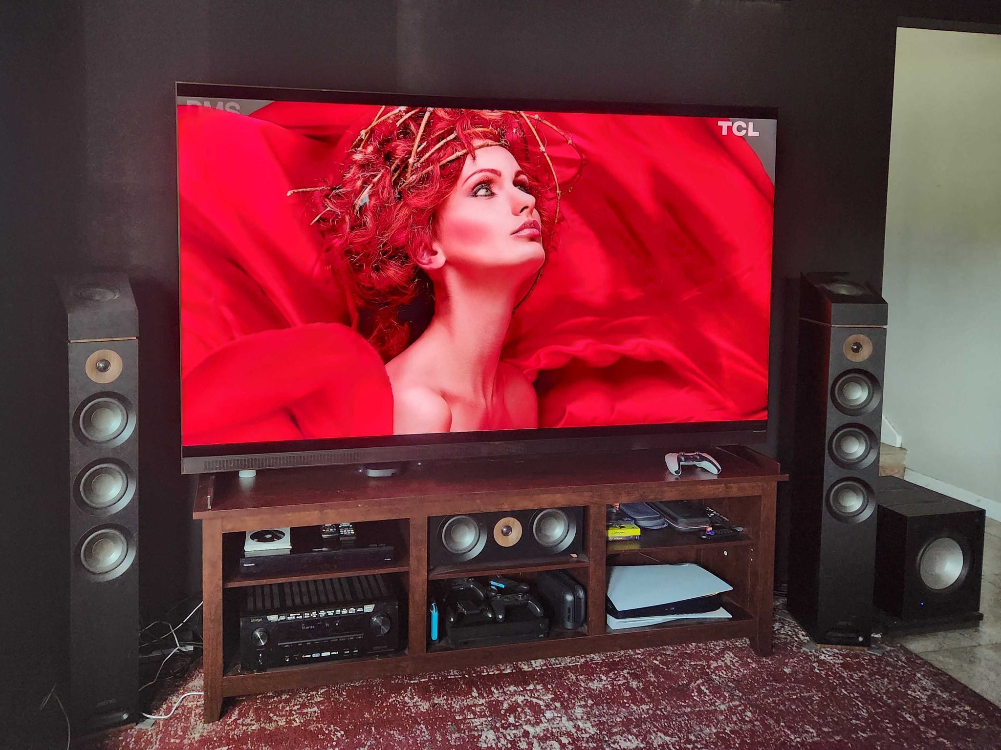 A TCL X11L TV on a media console in a living room with an image of a woman in red on screen.