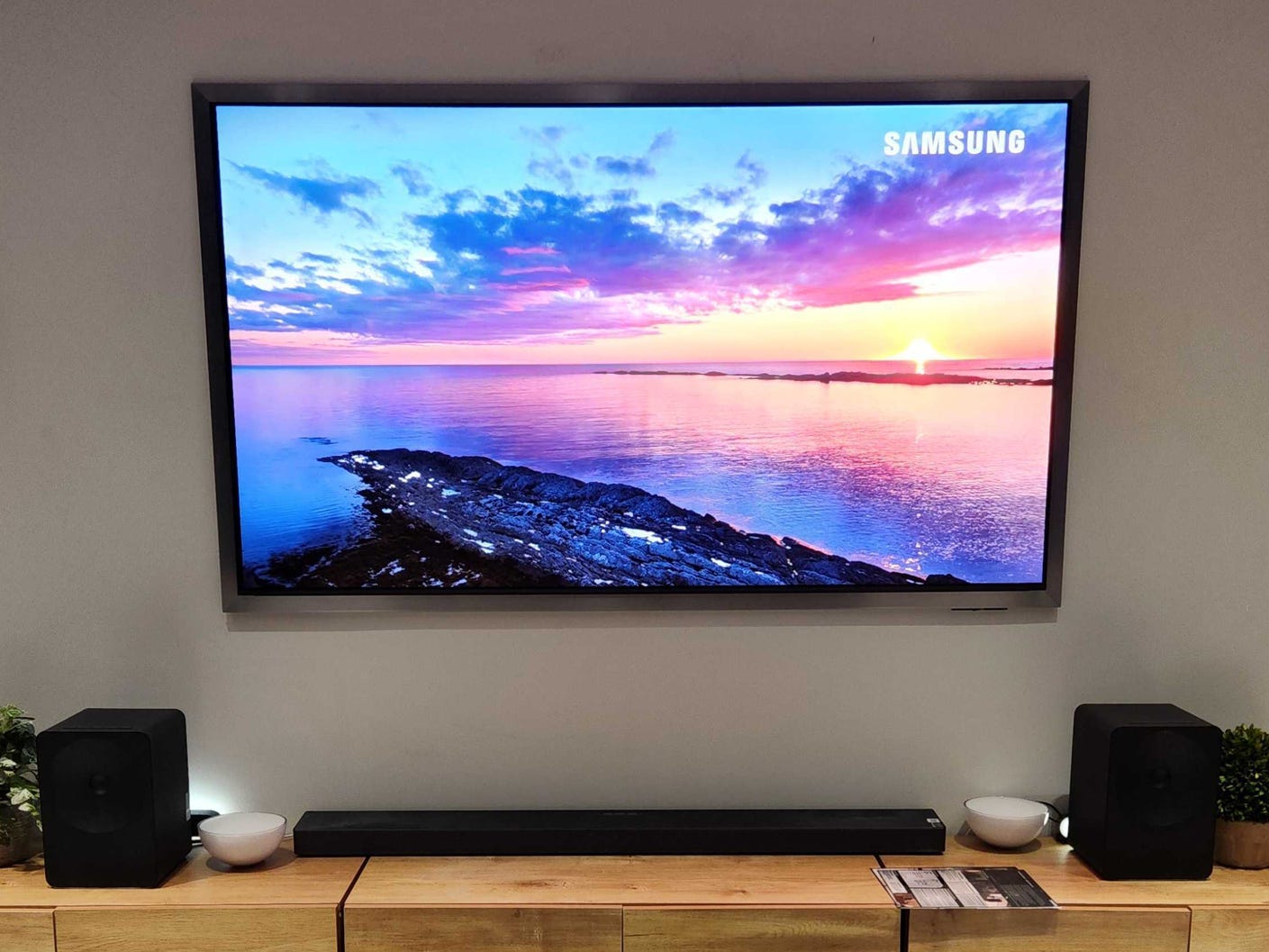 A Samsung S95H OLED TV hanging on a wall in a living room setup with an image of the ocean and a sunset on screen.