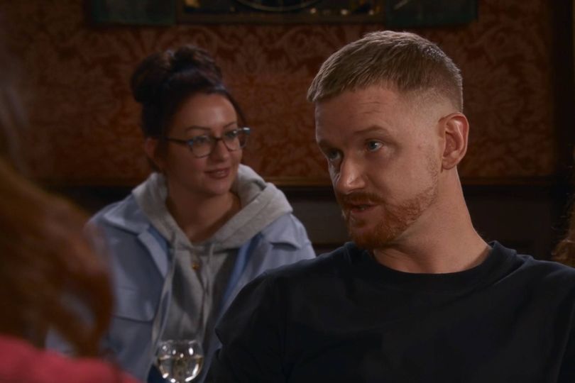 An indoor scene depicts two individuals engaged in a conversation. The person on the left, wearing glasses and a light-colored jacket, is looking towards the person on the right. The individual on the right, clad in a dark shirt, is focused on something or someone in front of them. The setting appears to be a casual indoor environment.