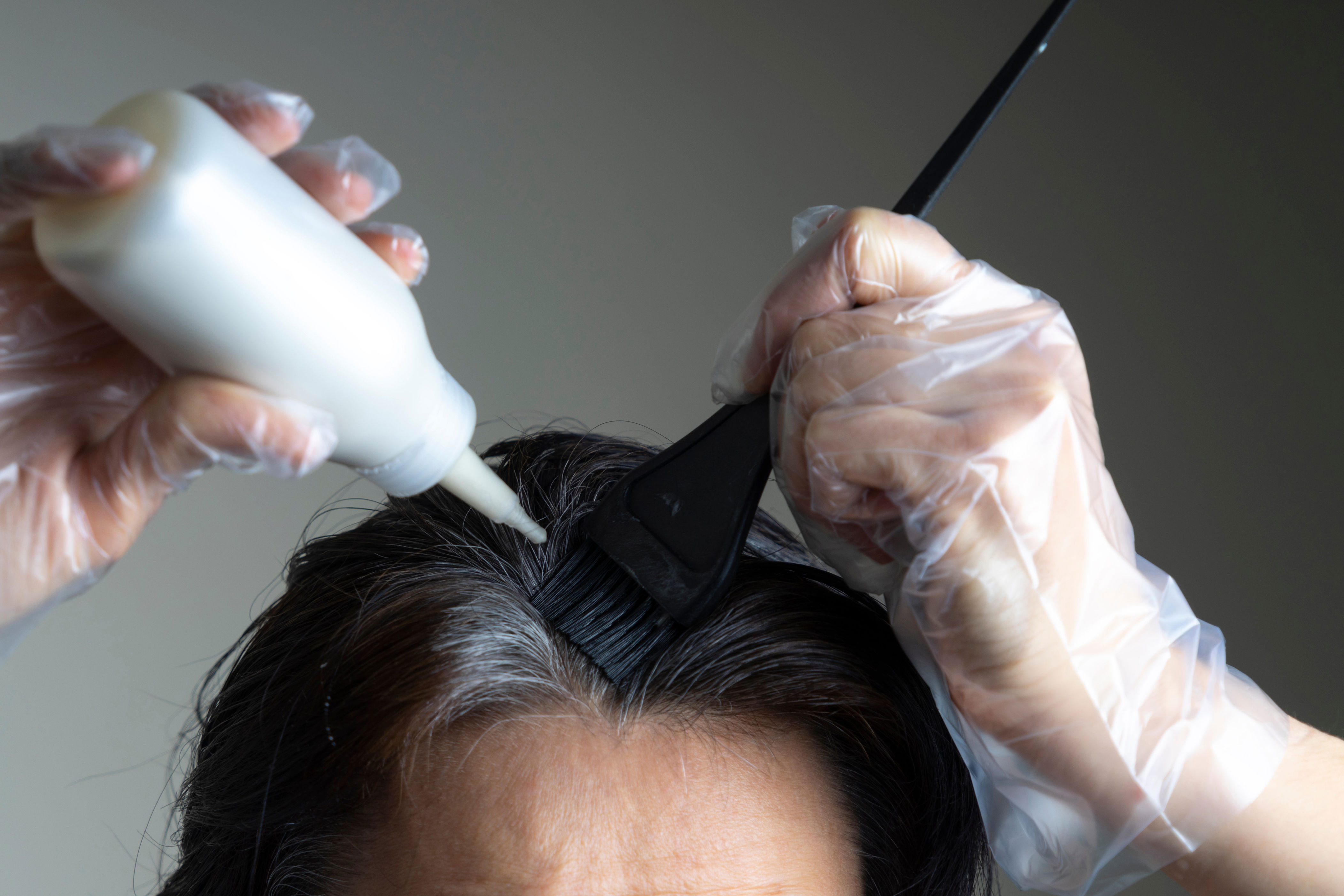 Person applying hair dye to their roots with a brush, wearing gloves and using a bottle applicator